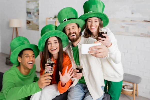 Positive bearded man taking selfie with interracial friends holding glasses of dark beer on Saint Patrick Day — Stock Photo