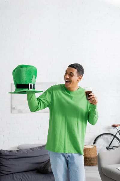 Happy african american man holding glass of dark beer while looking at green hat on Saint Patrick Day — Stock Photo