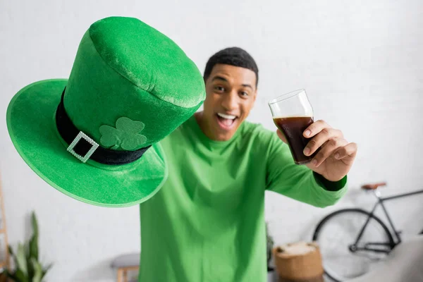 Happy african american man holding glass of dark beer and green hat on Saint Patrick Day — Stock Photo