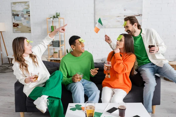 Woman pointing at bearded man with sticky note on forehead holding Irish flag while playing guess who game with interracial friends — Stockfoto