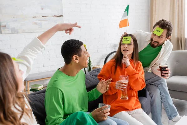 Женщина указывает на друзей с липкими нотами на лбу возле ирландского флага во время игры угадай, кто играет в день Святого Патрика — стоковое фото