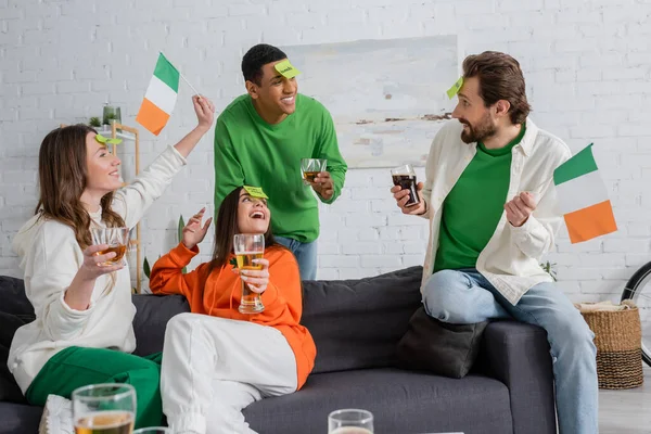 Щасливі міжрасові друзі з липкими нотатками на чолах, які тримають напої та ірландські прапори під час гри вгадуючи, хто грає на День святого Патрика. — стокове фото