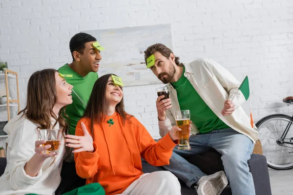 Cheerful multiethnic friends with sticky notes on foreheads holding alcohol drinks and playing guess who game on Saint Patrick Day — Stock Photo