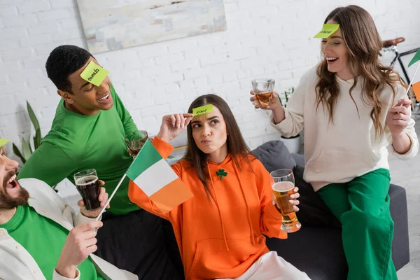 Multiethnic friends with sticky notes on foreheads holding alcohol drinks and looking at pensive woman playing guess who game — Stock Photo