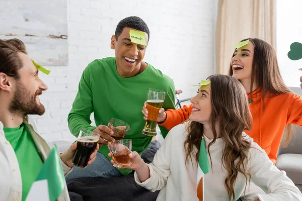 Happy multiethnic friends with sticky notes on foreheads clinking alcohol drinks and playing guess who game on Saint Patrick Day — Fotografia de Stock
