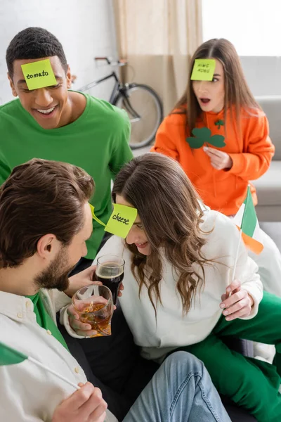 Улыбающиеся многонациональные друзья с липкими нотами на лбу, звенящие алкогольные напитки и играющие угадай, кто играет в день Святого Патрика — стоковое фото