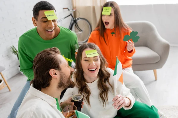Emotional interracial friends with sticky notes on foreheads holding alcohol drinks and playing guess who game on Saint Patrick Day — Stock Photo