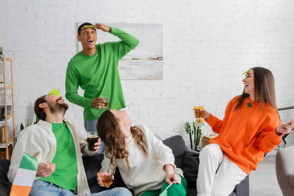Happy friends looking at african american man looking at sticky note on forehead while playing guess who game on Saint Patrick Day — Stock Photo