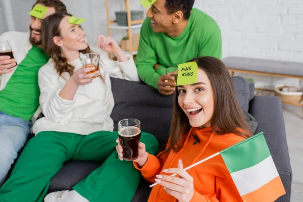 Взволнованная женщина с королем слово на липкой ноте с ирландским флагом и бокалом пива возле межрасовых друзей в День Святого Патрика — стоковое фото