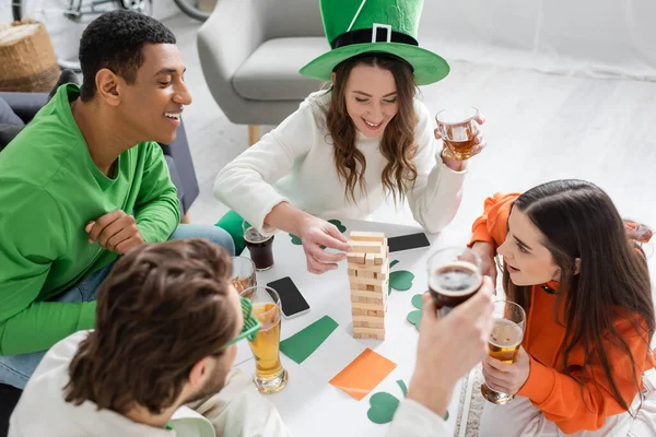 Overhead view of interracial friends with beer playing wood block game during saint patrick day at home — Stockfoto