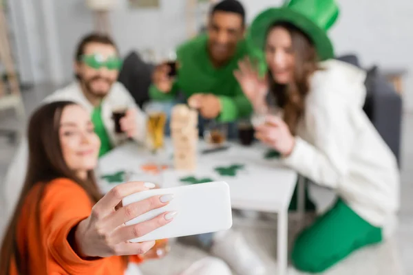 Blurred interracial friends taking selfie on smartphone while celebrating saint patrick day at home — Stockfoto