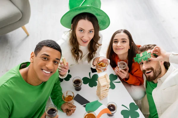 Overhead view of interracial friends celebrating saint patrick day and looking at camera at home — Stock Photo