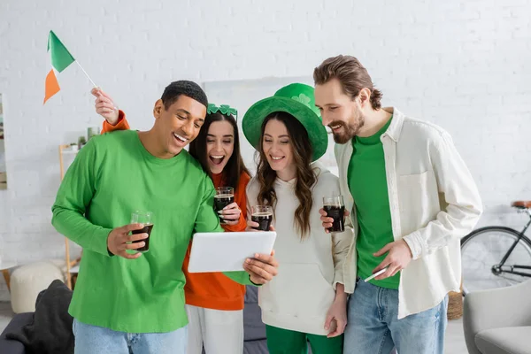 Позитивные межрасовые друзья с пивом с помощью цифрового планшета, празднуя День святого Патрика дома — стоковое фото