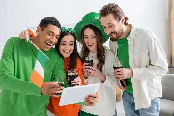 Позитивные многоэтнические друзья с пивом, празднуя День святого Патрика и используя цифровые планшеты дома — стоковое фото