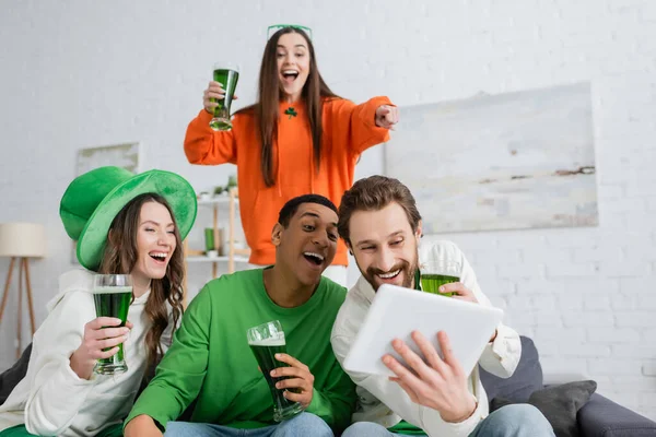Позитивные межрасовые друзья с зеленым пивом с помощью цифрового планшета во время Дня святого Патрика — стоковое фото