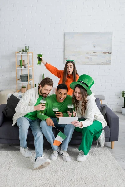Positive multiethnic friends with green beer using digital tablet on couch at home — Stock Photo