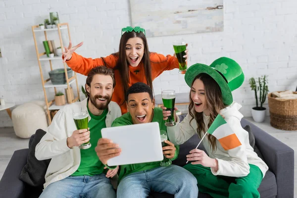 Emocionados amigos multiétnicos con bandera irlandesa y cerveza verde usando tableta digital en casa - foto de stock