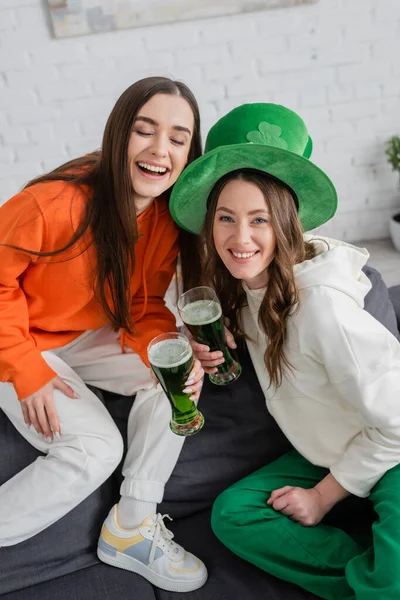 Mujeres jóvenes positivas sosteniendo cerveza verde durante la celebración de San Patricio en casa - foto de stock