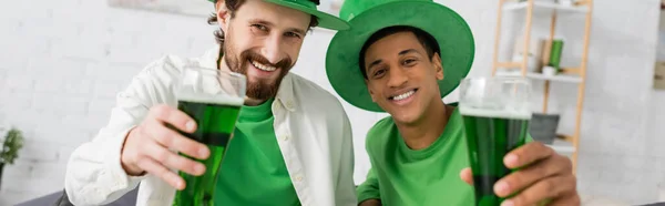 Cheerful interracial friends holding green beer during saint patrick day, banner — Stock Photo