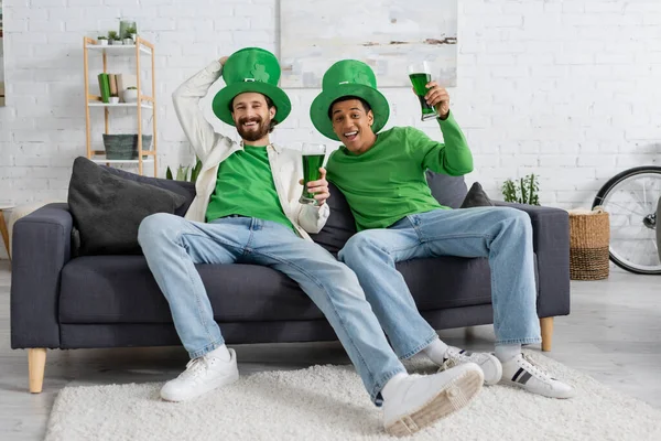 Улыбающиеся многонациональные друзья в праздничных шляпах, держащие зеленое пиво во время празднования дня святого Патрика — стоковое фото