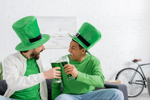 Excited multiethnic friends in green hats clinking beer during saint patrick day at home — Photo de stock