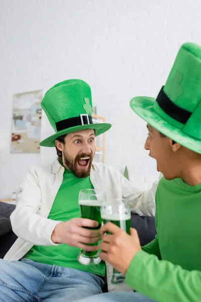 Захоплений чоловік в зеленому капелюсі тримає пиво біля афроамериканського друга вдома — стокове фото