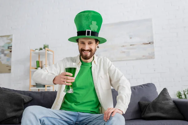 Hombre sonriente con sombrero verde sosteniendo cerveza y mirando a la cámara durante la celebración de San Patricio en casa - foto de stock