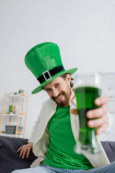 Hombre sonriente con sombrero sosteniendo cerveza verde borrosa durante la celebración de San Patricio en casa - foto de stock