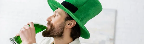 Seitenansicht eines bärtigen Mannes mit Hut, der zu Hause grünes Bier trinkt, Banner — Stockfoto