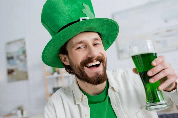 Alegre hombre en santo patrick sombrero sosteniendo cerveza verde y mirando a la cámara en casa - foto de stock