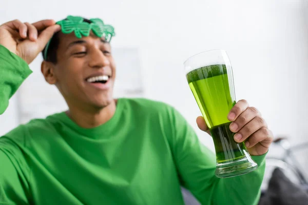 Homem americano africano desfocado segurando óculos escuros e cerveja verde em casa — Fotografia de Stock
