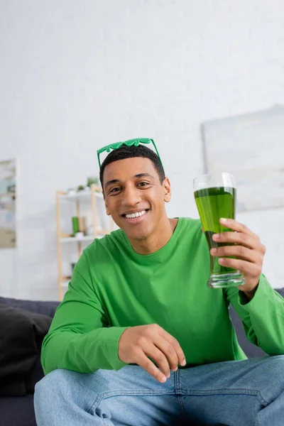 Cheerful african american man holding green beer and looking at camera at home — Stock Photo
