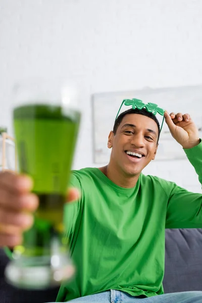 Позитивный африканский американец, держащий очки для вечеринок и размытое пиво дома — стоковое фото