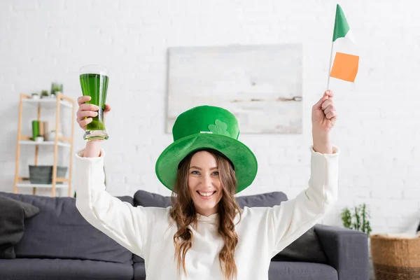Jovem alegre de chapéu verde segurando bandeira irlandesa e cerveja em casa — Fotografia de Stock