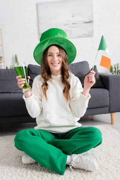 Jovem mulher positiva de chapéu segurando cerveja verde e bandeira irlandesa em casa — Fotografia de Stock