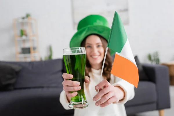 Mulher desfocada de chapéu segurando cerveja verde e bandeira irlandesa em casa — Fotografia de Stock