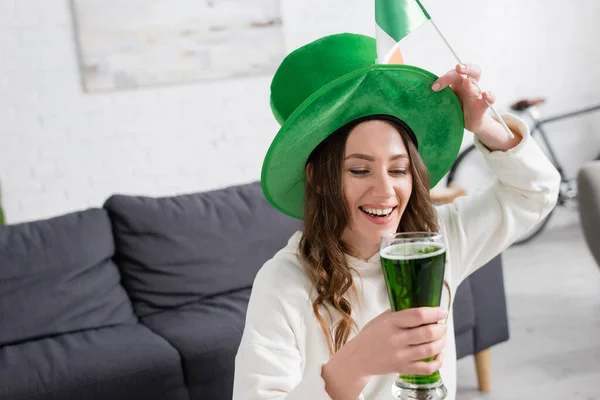 Mulher alegre de chapéu segurando cerveja verde e bandeira irlandesa em casa — Fotografia de Stock