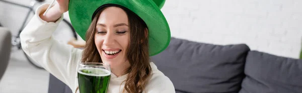 Positive young woman in hat holding glass of green beer at home, banner — Stock Photo