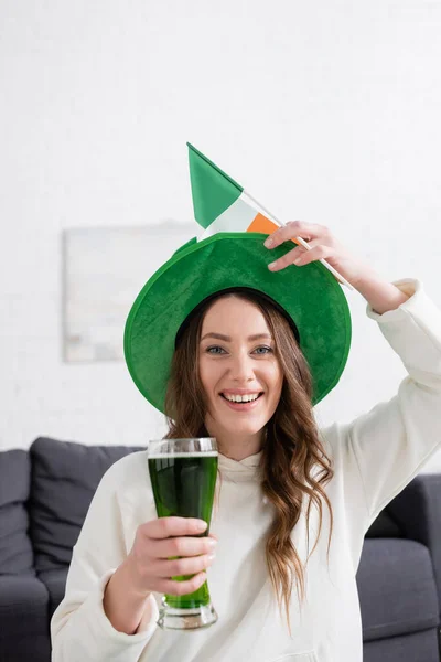 Positive woman in hat holding green beer and Irish flag while looking at camera at home — Photo de stock