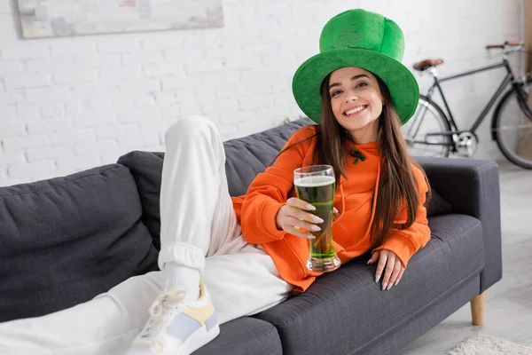 Sorridente giovane donna in cappello verde che tiene la birra mentre giace sul divano — Foto stock