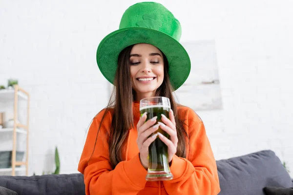 Улыбающаяся женщина в шляпе святого Патрика Дэя держит пиво в гостиной — стоковое фото