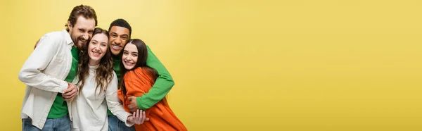 Smiling interracial friends hugging and looking at camera isolated on yellow, banner — Stock Photo