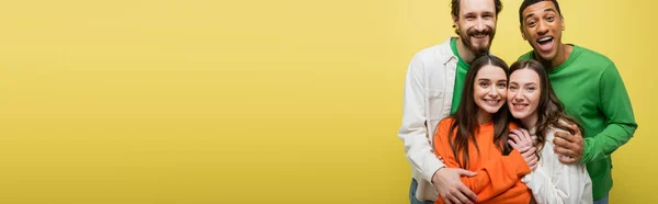 Excited interracial men hugging young women isolated on yellow, banner — Stock Photo