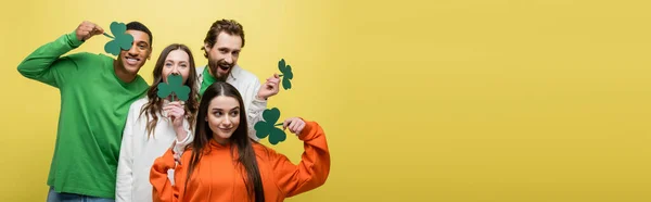 Positive interracial friends holding green paper clover isolated on yellow, banner — Stock Photo