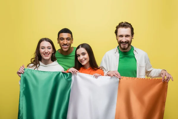 Позитивные мультиэтнические люди держат ирландский флаг изолированным на желтом — стоковое фото