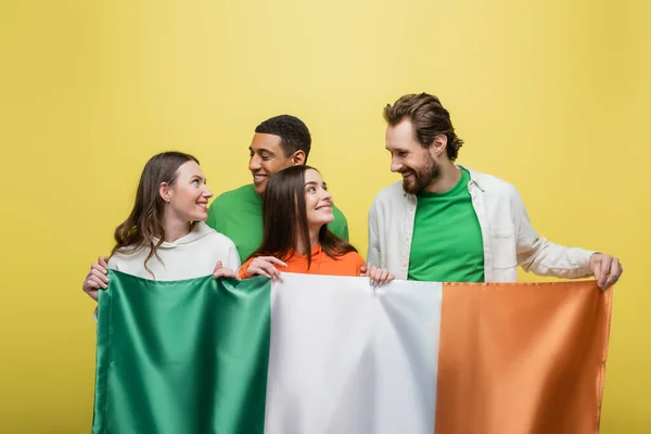 Interracial amigos olhando uns para os outros enquanto segurando bandeira irlandesa isolado no amarelo — Fotografia de Stock