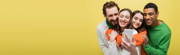 Cheerful woman taking selfie with interracial friends isolated on yellow, banner — Stock Photo