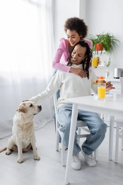 Smiling african american woman hugging boyfriend near labrador and breakfast in kitchen - foto de stock
