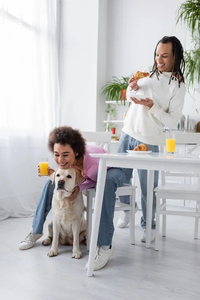 Positive african american woman hugging labrador near boyfriend with croissant in kitchen — Stock Photo