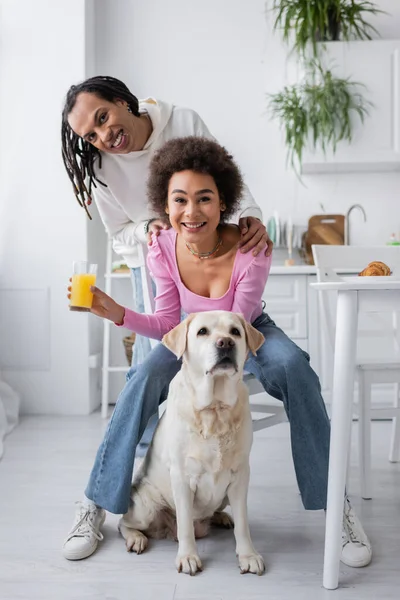 Sonriente pareja afroamericana mirando a cámara cerca de labrador en cocina por la mañana - foto de stock